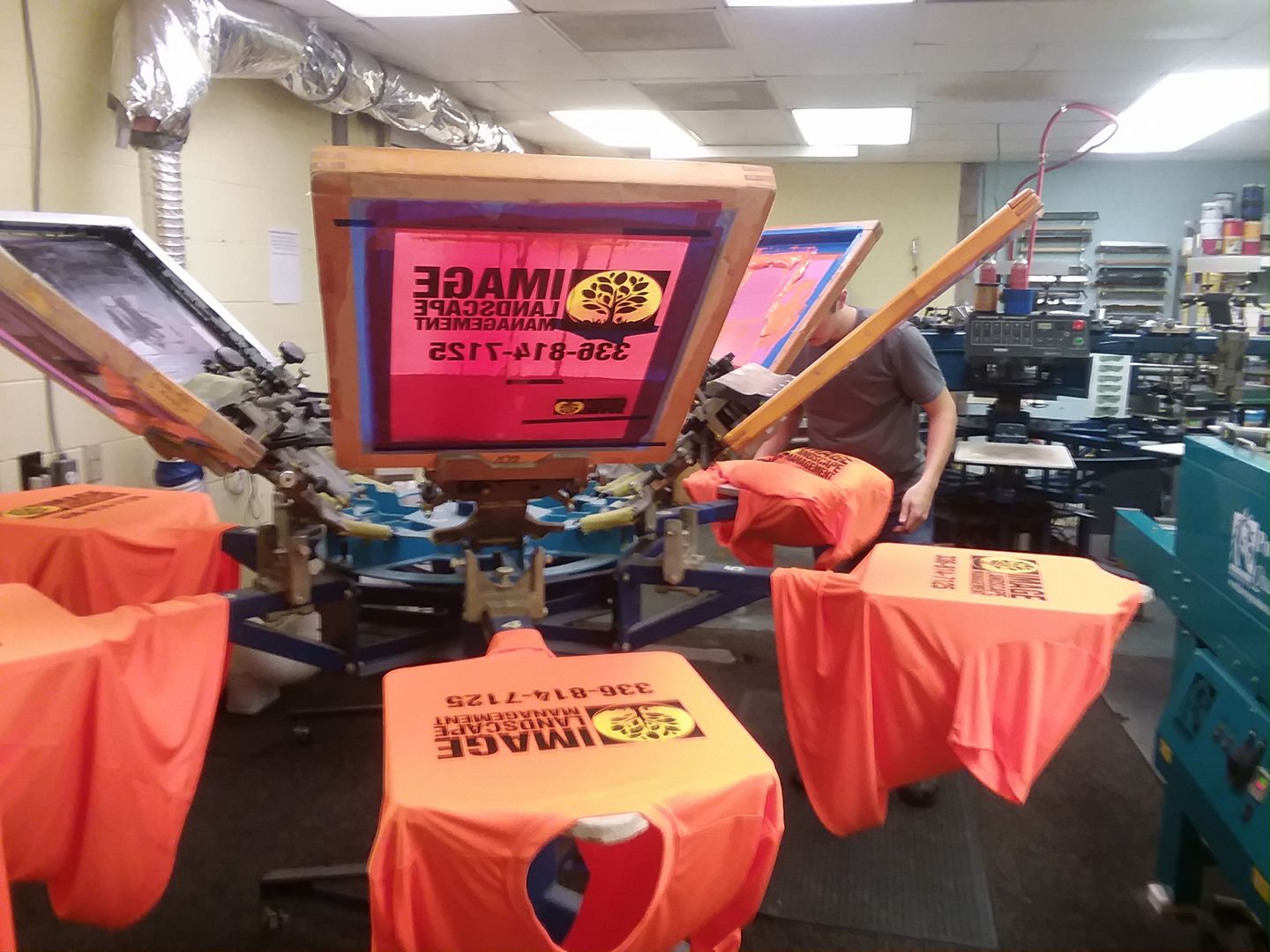 A person operates a screen printing machine to print orange T-shirts in a workshop.